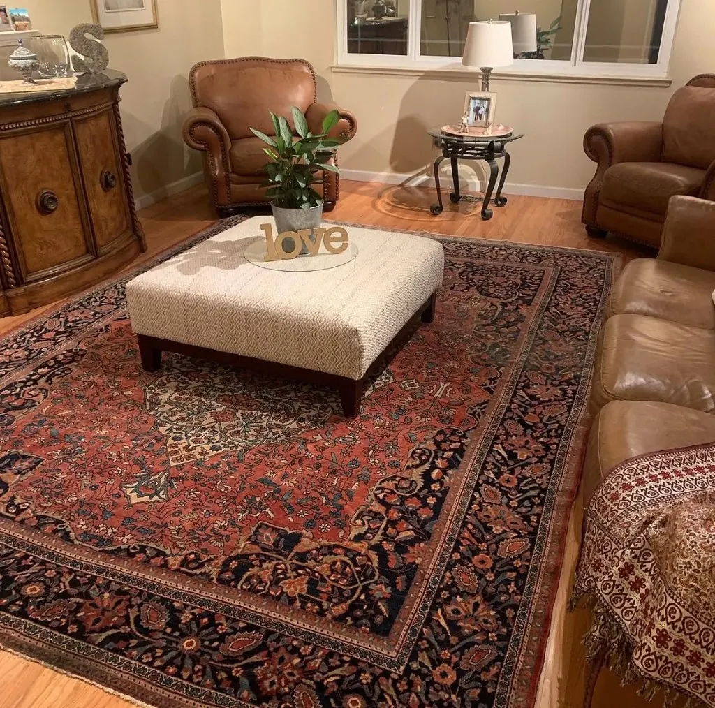 Living room anchored by a large Persian-style rug in terracotta, navy, and cream, with leather seating, a central ottoman, and warm ambient light